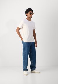 T-shirt scurt cu mânecă, de culoare bej deschis, blugi de denim albastri și adidași albi. Modelul stă pe un fundal simplu, purtând ochelari de soare maro.