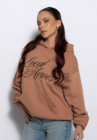 Brązowa bluza z kapturem z napisem "Local Heroes" w czerni, z przednią kieszenią i długimi rękawami. Połączona z jasnoniebieskimi dżinsami. Gładka tekstura.