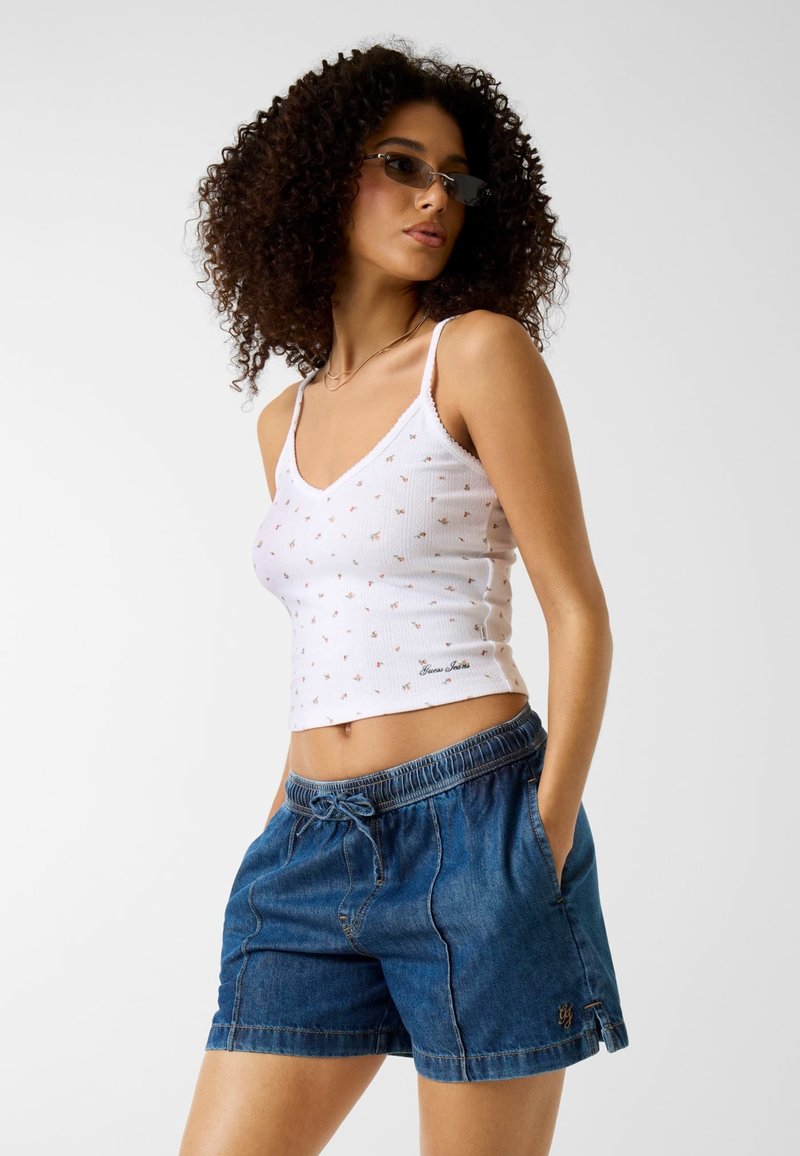 Göndör hajú nő, kicsi napszemüveget visel, fehér virágmintás ujjatlan felsőt és kék farmer rövidnadrágot, kezét a zsebében tartva pózol fehér háttér előtt.