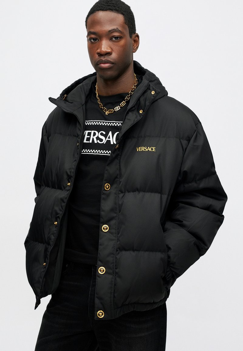 Sort puffjakke med hætte, der har gyldent hardware, lynlås og "VERSACE" logo i guld på brystet. Båret over en sort skjorte.