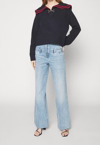 Marinblå tröja med röda ränder på kragen och guldiga detaljer, kombinerad med ljusblå utsvängda jeans och svarta sandaler med remmar.