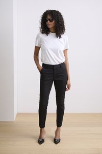 Pantalons noirs ajustés, t-shirt blanc à manches courtes et escarpins noirs à bout pointu. Modèle portant des lunettes de soleil sombres avec des cheveux bouclés. Fond minimaliste.