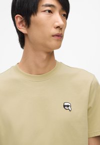 Olivovo zelené bavlnené tričko s okrúhlym výstrihom a čiernobielou vyšitou nášivkou na ľavej hrudi znázorňujúcou postavu so slnečnými okuliarmi.