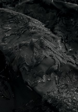 Schwarzer Stoff mit texturiertem Stickmuster und fransigen Kanten sowie dezenten metallischen Fäden für zusätzliche Details. Glatte darunterliegende Schicht sichtbar.