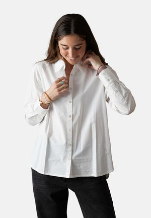 Mujer con cabello largo y castaño que lleva una camisa blanca abotonada y pantalones negros, ajustándose el cuello y sonriendo mientras mira hacia abajo.