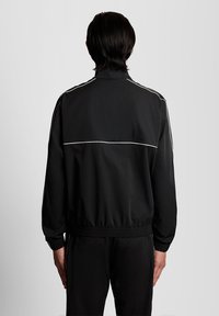 Άτομο που στέκεται με την πλάτη προς την κάμερα, φοράει μαύρο αθλητικό μπουφάν και παντελόνι με λευκές ρίγες στα μανίκια και στην πάνω πλάτη.