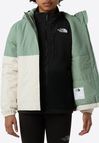 Veste color block verte, crème et noire avec capuche, dotée d'une doublure intérieure en polaire et d'un logo de marque sur la poitrine.