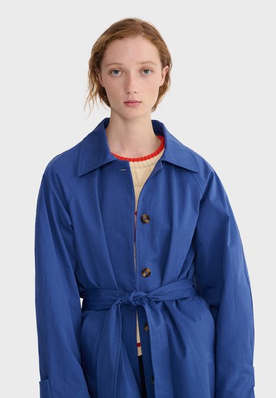 Jeune personne à la peau claire et aux cheveux roux portant un manteau bleu ceinturé sur un pull crème avec une bordure rouge, face à un fond uni.