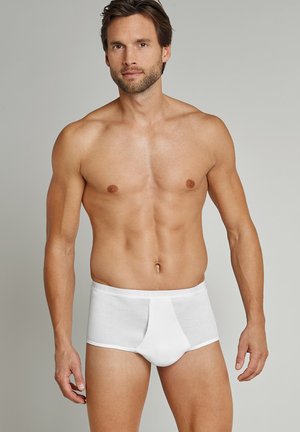 Uomo senza camicia che indossa slip bianchi a coste, in piedi di fronte a uno sfondo grigio uniforme, con le braccia rilassate.