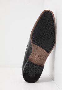 Suola di scarpa nera lucida con gomma nera testurizzata e accenti marroni, caratterizzata da un'impronta del logo e un tacco leggermente rialzato.