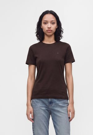 Femme aux cheveux bouclés mi-longs portant un t-shirt à manches courtes marron foncé et un jean bleu, debout devant un fond blanc uni.