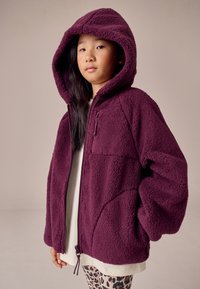 Vinrød fleecejakke med stor hætte, lynlås, to forlommer og en afslappet pasform, båret over en lys skjorte og mønstrede leggings.