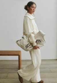 Felpa beige oversize e pantaloni abbinati, accompagnati da una grande borsa a tracolla crema con scritte marroni. Panchina in legno e sfondo neutro.