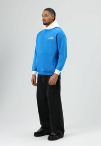 Blauwe trui met een witte kap, voorzak en logo detail. Gecombineerd met zwarte wijde broekspijpen en zwarte schoenen met roze veters.