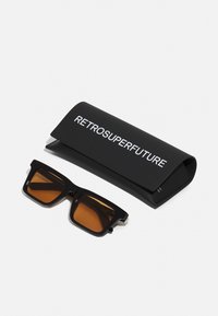 Musta retro stiilis päikeseprillid pruuni objektiiviga, esitatud koos sobiva musta karbiga, millel on valge tekst "RETROSUPERFUTURE".