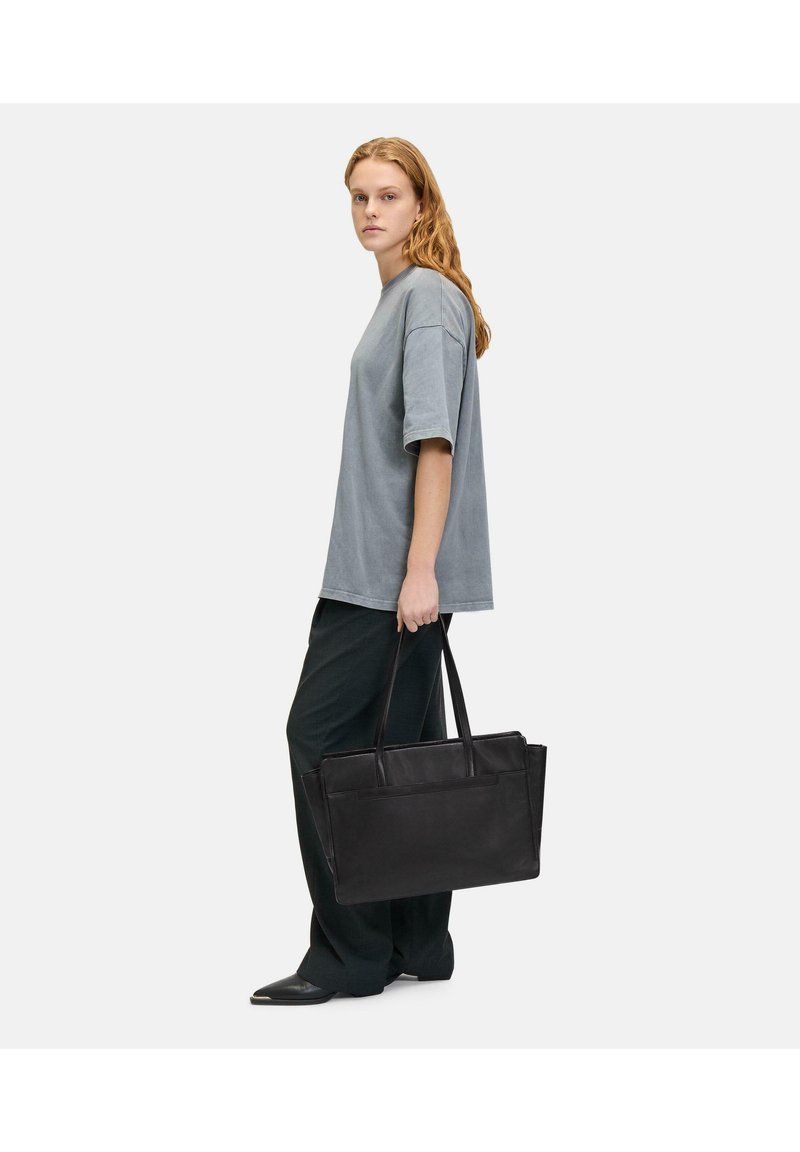 Czarna skórzana torebka typu tote z podwójnymi uchwytami, prostokątnym kształtem i eleganckim designem. Modelka ma na sobie szary, oversize'owy t-shirt oraz ciemne spodnie.
