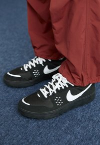 Sapatilhas Nike pretas com acabamentos brancos, parte superior em malha e bico redondo; combinadas com calças vermelhas de corte largo, sobre um tapete azul.