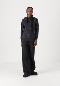 Sudadera negra con bordado de estrella blanca, capucha con cordón, mangas largas, combinada con jeans negros de pierna ancha y zapatos negros gruesos.