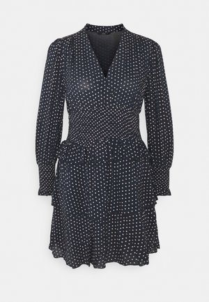 Granatowa sukienka z długimi rękawami, dekoltem w kształcie litery V i elastycznym paskiem w talii. Posiada falbaniaste wykończenie i wzór w białe kropki. Lekki, zwiewny materiał.