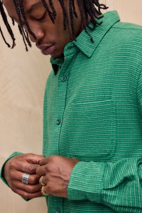 Grønternet skjorte med knapper og en lomme, fremstillet i et tekstureret stof med kontrastfyldte blå knapper. Hænder med ringe på.