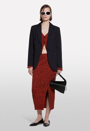 Blazer noir sur un crop top texturé rouge et une jupe assortie avec une fente. Le mannequin tient un sac structuré noir, porte des lunettes de soleil blanches et des ballerines noires.