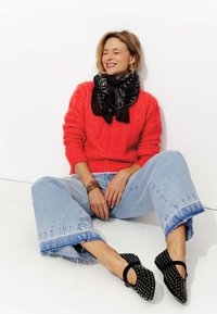 Pull en tricot rouge, écharpe noire à motifs, jeans taille haute bleu clair avec ourlet effiloché et ballerines noires à clous. Les accessoires incluent des bracelets.