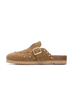 Zueco de ante marrón con tachuelas decorativas y un detalle de hebilla. Cuenta con una suela contorneada con borde dentado para mayor agarre y una textura suave.