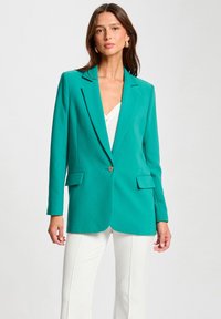 Blazer entallado en color verde azulado con solapas de muesca, botón frontal único y bolsillos frontales. Estilizado con una blusa blanca y pantalones de color claro.