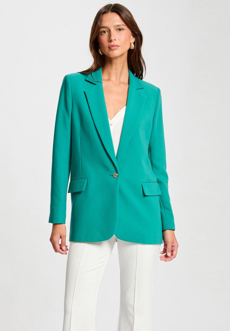 Blazer entallado en color verde azulado con solapas de muesca, botón frontal único y bolsillos frontales. Estilizado con una blusa blanca y pantalones de color claro.