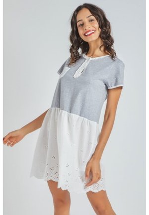 Mujer sonriente con cabello rizado que lleva un vestido gris de manga corta con falda de encaje blanco con ojales y borde ondulado, posando sobre un fondo claro.