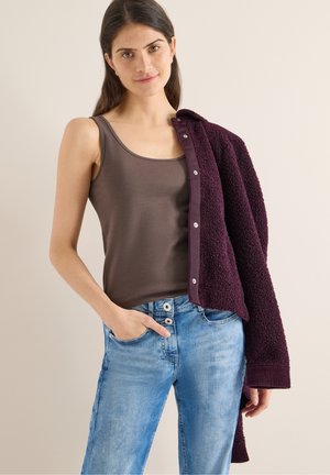 Brązowy top na ramiączkach, ciemnofioletowy teksturowany kardigan, niebieskie dżinsy z dwoma guzikami. Modelka pozuje z jedną ręką w kieszeni, a kardigan zarzucony na ramię.