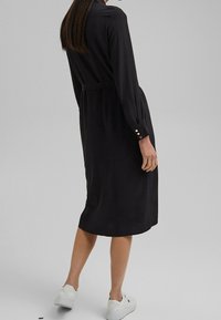 Czarna, długorękawowa sukienka z paskiem w talii, z guzikami przy mankietach i gładką strukturą, sięgająca do połowy łydki, zestawiona z białymi trampkami.
