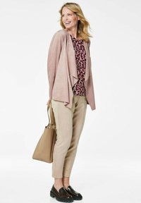 Hellrosa, aus Kunstwildleder gefertigte offene Strickjacke, mit einem gemusterten burgunderrot- und cremefarbenen Oberteil, hellbeigen, schmal zulaufenden Hosen und dunkelbraunen Slippern, sowie einer beigen Tragetasche.