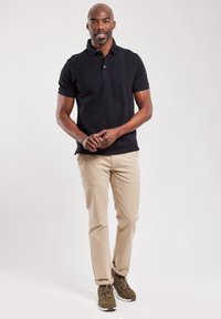 Zwarte polo met korte mouwen en een kraag, gecombineerd met beige chino's en olijfgroene sneakers, staand tegen een lichte achtergrond.