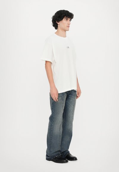 T-shirt blanc à manches courtes avec un petit graphique sur la poitrine, associé à un jean bleu clair évasé et des chaussures noires. Tissu lisse, coupe décontractée.