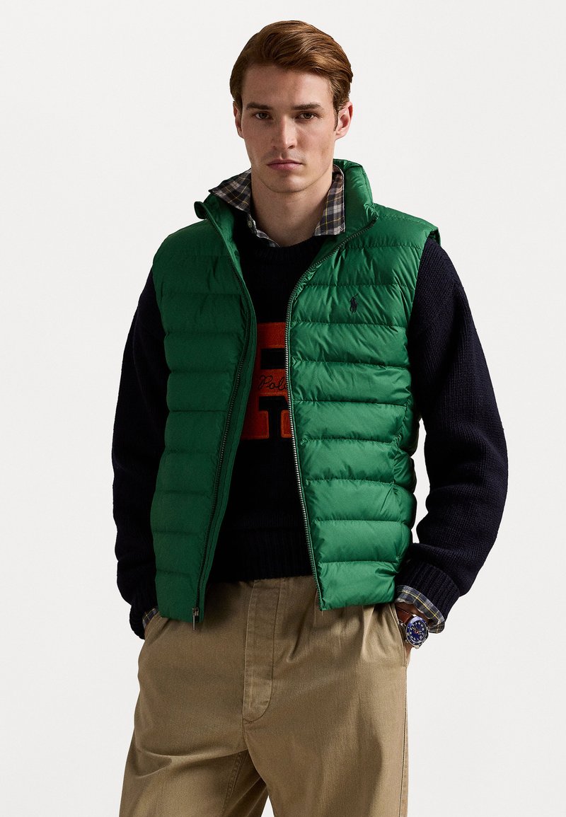 Green puffer vest with quilted panels, zip closure, and high collar. Worn over a navy sweater and plaid shirt, paired with beige pants.