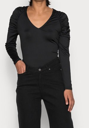 Personne portant un top noir à col en V avec des manches longues froncées et un jean noir taille haute, une main dans la poche sur un fond uni.