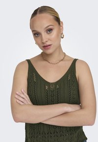 Top sin mangas de punto verde con escote en V y textura de encaje calado. El modelo muestra los brazos cruzados, con pendientes de aro dorados y collar de cadena.