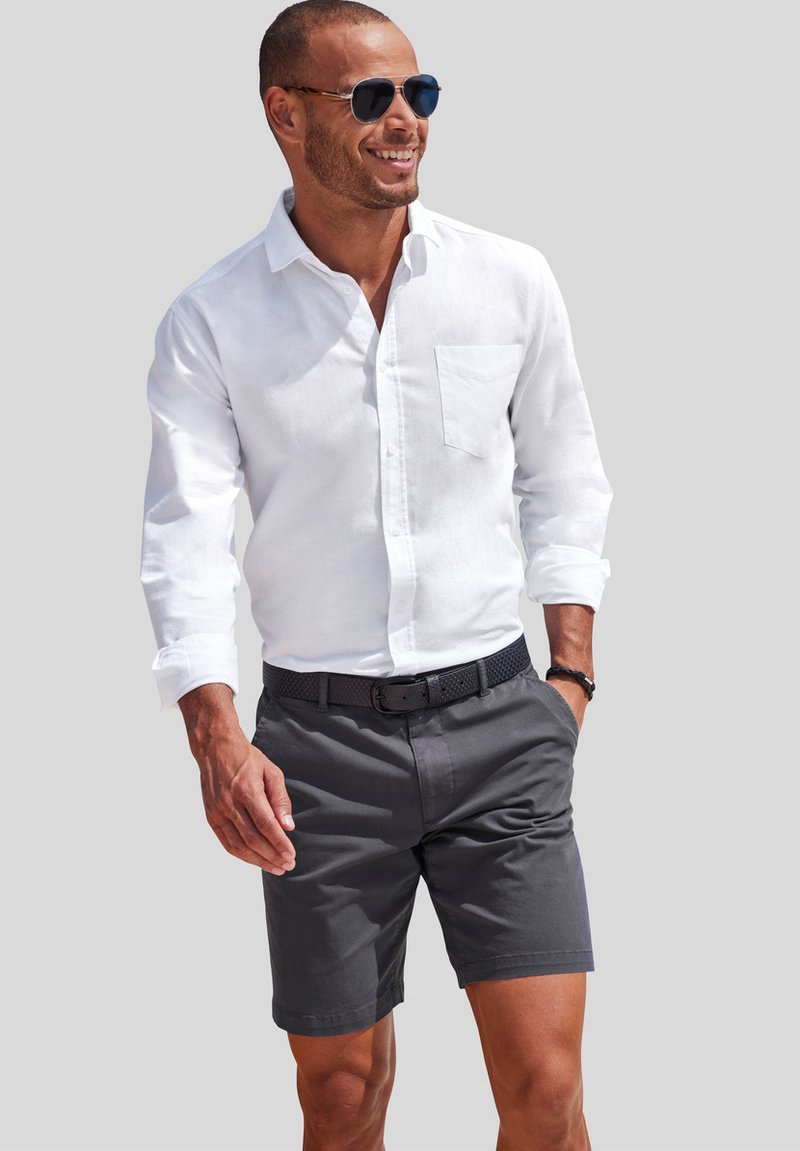 Camicia bianca a maniche lunghe con colletto a bottoni, pantaloni corti grigi, cintura nera texture e occhiali da sole, posizionata su uno sfondo neutro.