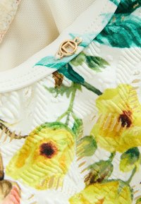 Witte stof met levendige gele en groene bloemenpatronen. Een gouden ronde sluiting is zichtbaar nabij de halslijn. Textuurdetail.