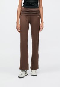 Καφέ παντελόνι φαρδύ με σούρες στη μέση, κατασκευασμένο από μαλακό ύφασμα, με μικρό λογότυπο Nike στο ισχίο. Φορώντας το με γκρι αθλητικά παπούτσια.