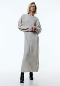 Langes, beige gestricktes Kleid mit Ballonärmeln und einer Naht in der Mitte. Kombiniert mit schwarzen Knöchelstiefeln, die eine elegante Silhouette schaffen.