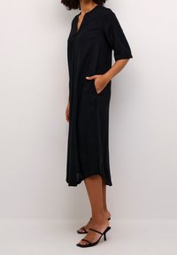 Vestido negro de longitud hasta la rodilla con escote en V, mangas cortas y bolsillos laterales. Tejido suave con un corte relajado y silueta alargada.