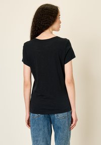 Moteris su garbanotais plaukais, dėvinti paprastą juodą trumpomis rankovėmis marškinėlį ir mėlynas džinsus, atsukusi nugarą priešais šviesų paprastą foną.