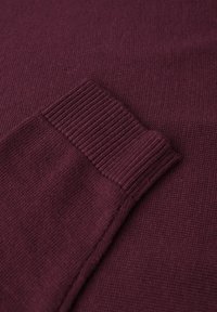 Maglione bordeaux con una morbida trama a maglia, con polsini a coste e un corpo liscio. La vista ravvicinata evidenzia le cuciture dettagliate.