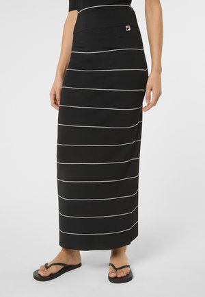 Kobieta ubrana w czarną maxi spódnicę z cienkimi białymi poziomymi paskami oraz czarne klapki, stojąca na jednolitym tle.