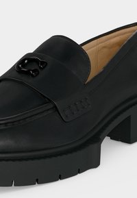 Gros plan sur un mocassin en cuir noir avec une semelle épaisse, un détail métallique décoratif sur le devant et une doublure intérieure beige.