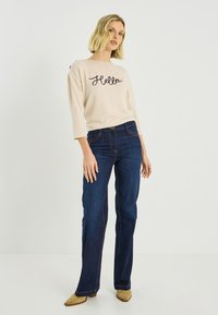 Top beige de manga larga con bordado "Hello", combinado con jeans azul oscuro. La modelo lleva tacones beige de punta puntiaguda. Conjunto sencillo y casual.