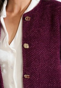 Gros plan sur une blouse blanche sous une veste texturée violette foncée avec trois boutons en or de forme irrégulière, montrant une partie du cou et de la clavicule d'une personne.