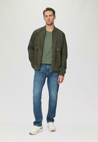 Oliwkowa kurtka bomberka z dwoma przednimi kieszeniami, zestawiona z dopasowanym zielonym t-shirtem i niebieskimi dżinsami. Białe sneakersy dopełniają stylizację.